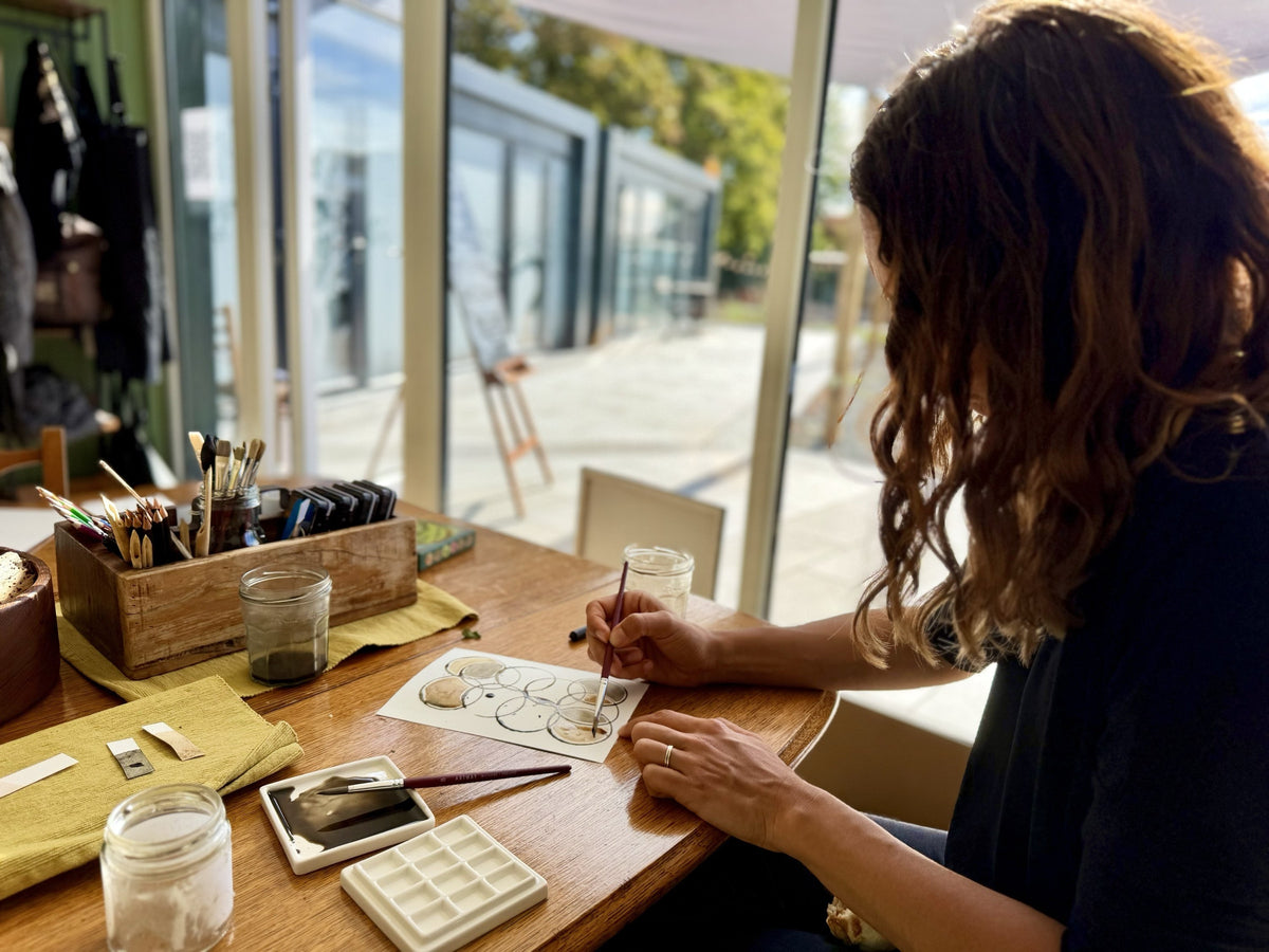 Louise painting ink circles at the studio table, with brushes, jars of ink and the glass front looking out to the farm